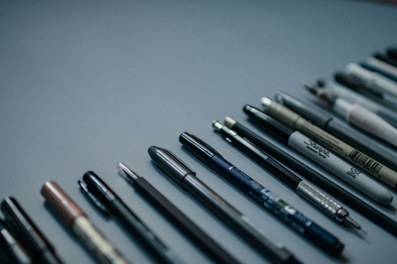 A row of assorted pens laid diagonally on a grey surface
